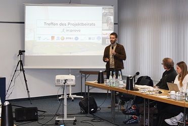 Das Bild zeigt einem Raum, in dem ein Mann mit Mikrofon neben einer Leinwand einen Vortrag hält und ihm Menschen zuhören (verweist auf: in:prove: Gewinn­bringender Austausch des Projekt­beirats in Bonn)